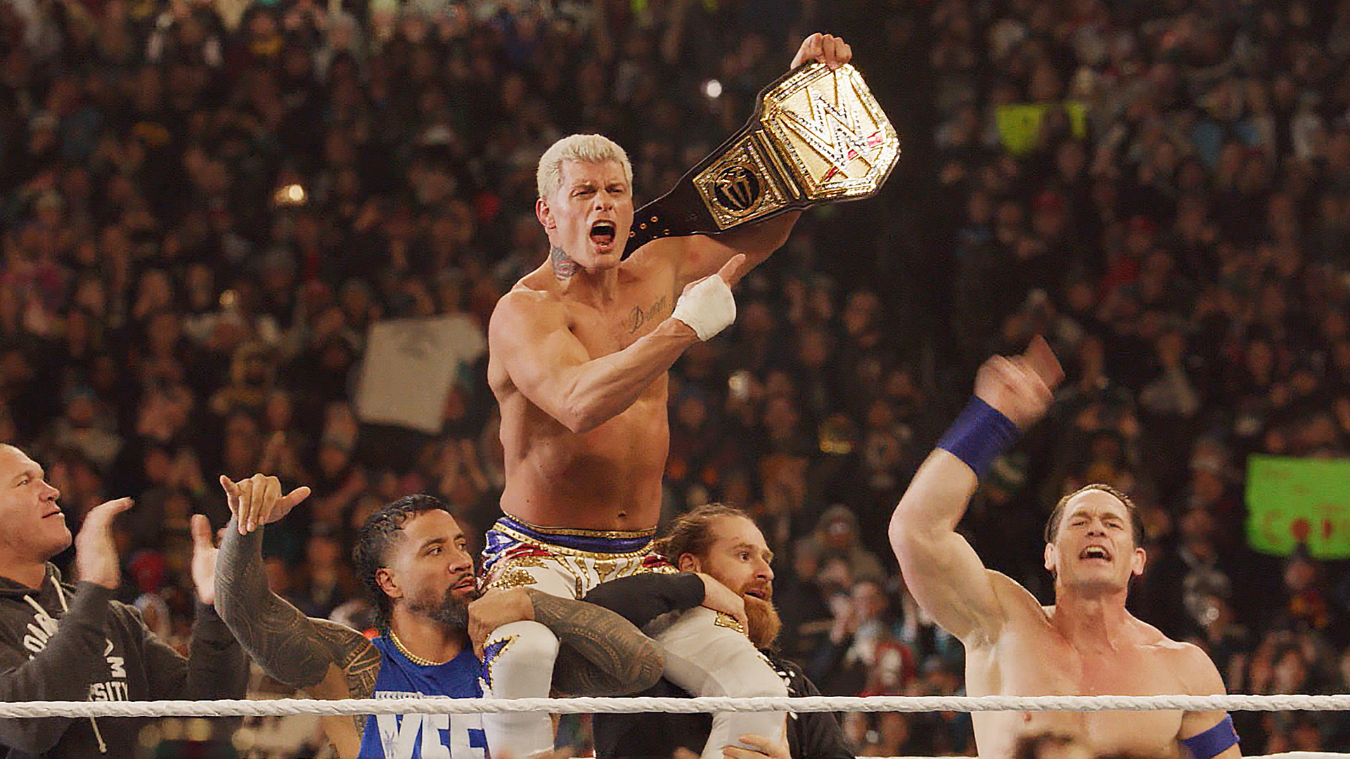 Cody Rhodes on the shoulders of other wrestlers as he points to his belt