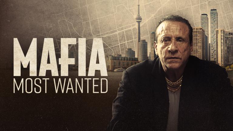 A stern man in a dark suit and gold chain stands before a city skyline and map background. Text reads “Mafia: Most Wanted.”